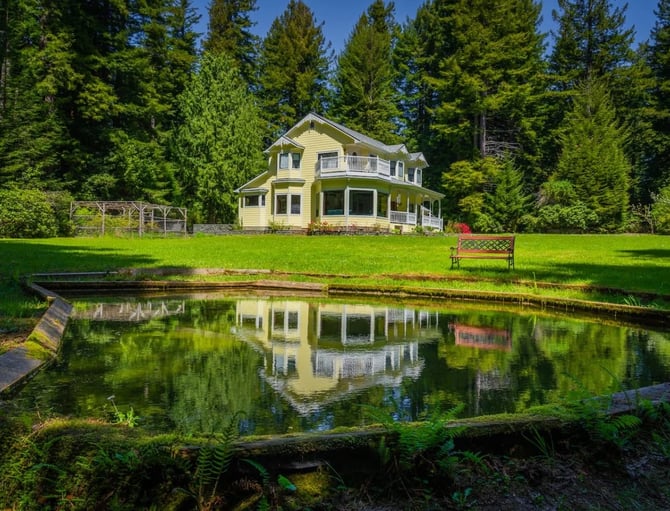 Yellow Victorian house with wraparound porch surrounded by tall evergreen trees, green lawn, and small pond with reflection in foreground.